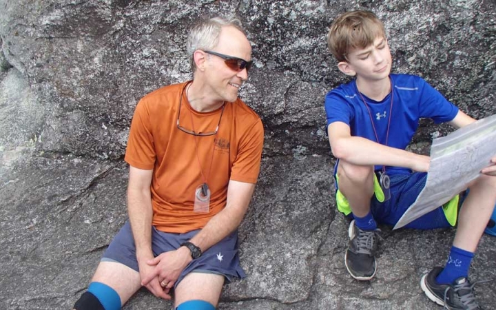 A parent and child rest on a rock and examine a map the child is holding and pointing too.